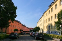 Straße mit Kopfsteinpflaster und Autos, links Häuser in orange, rechts Häuser in gelb.