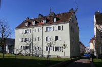 Altbau mit Spitzdach, 4 Etagen, hellgrüne Fassade. Vorne Grünfläche, Wäschestangen. Im Hintergrund Straße.