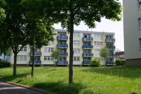 Flachdachgebäude. 4 Etagen, Fassade hellgelb, Balkone blau. Davor Wiese und Bäume.