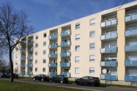 Flachdachgebäude mit fünf Etagen, Loggien. Hellgelbe Fassade. Davor Grünstreifen und Straße mit parkenden Autos.