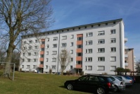 Hellgraues Gebäude mit fünf Wohnetagen, Schrägdach, davor eine Wiese.