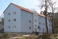 Saniertes, hellblaues Mehrfamilienhaus mit Satteldach, 3 Etagen. Rechts Bäume im Winter bei Sonnenschein.