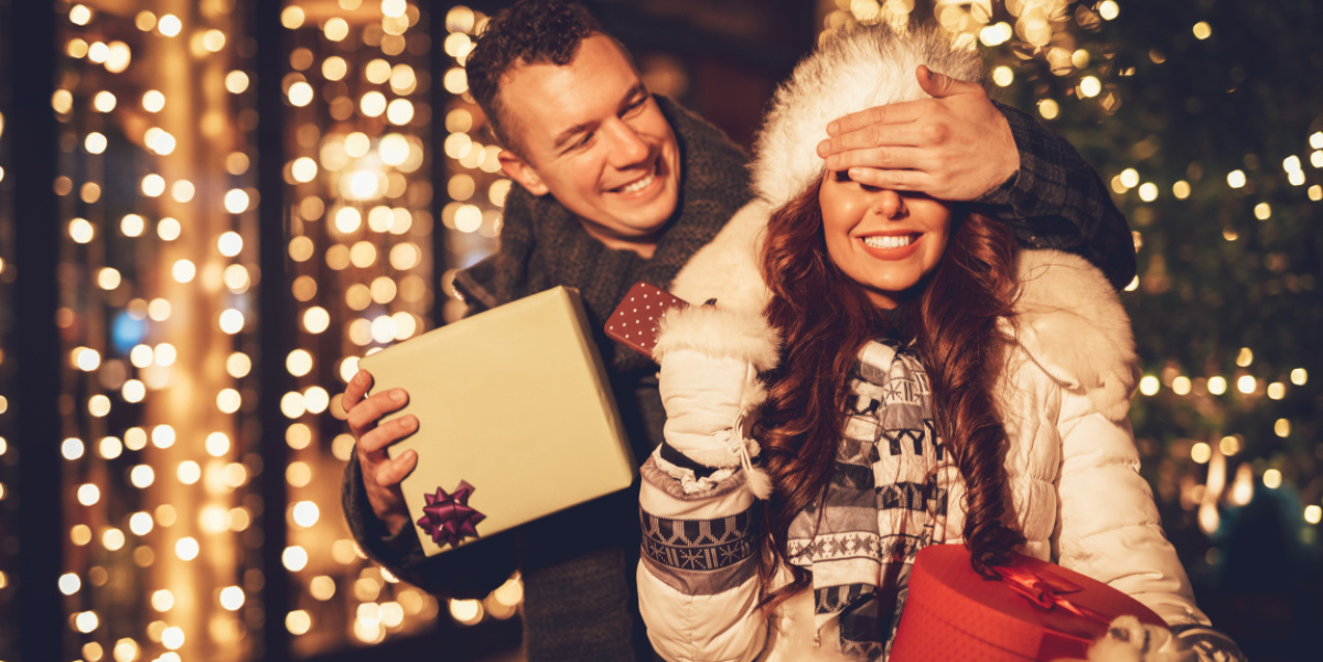 Schenken im Advent Ein Paar vor einem weihnachtlich beleuchtetem Schaufenster. Man hält Frau die Augen zu, um sie mit einem Geschenk zu überraschen.