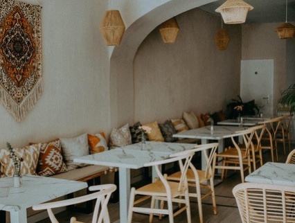 Innenraum von Café Batela mit  weißen Tischen und Rattanstühlen mit Kissen und Wanddekoration im Boho-Stil