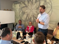 Eine Person steht, um sie herum sitzen mehrere Personen an einem Tisch. 