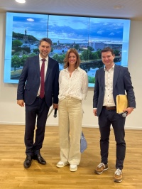 Links  steht ein Mann im Anzug mit Mikrofon, in der Mitte eine Frau mit Tüte, rechts ein Mann mit einem Umschlag und Mikrofon