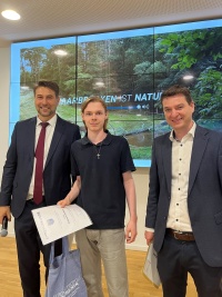 Links  steht ein Mann im Anzug mit Mikrofon, in der Mitte ein junger Mann mit Urkunde und Tüte, rechts ein Mann im Anzug