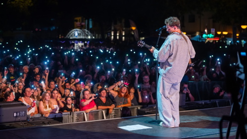 Ein Sänger steht auf einer Bühne im Scheinwerferlicht. Er hält eine Gitarre, trägt lockere Kleidung und eine Brille. Vor ihm ist ein Mikrofon auf einem Ständer. I