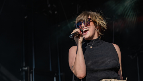 Sängerin singt leidenschaftlich mit Sonnenbrille und schwarzem Top vor dunklem Bühnenhintergrund.