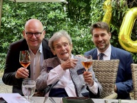 Eine alte Dame mit einem Glas sitzt an einem Tisch, dahinter zwei Männer in gebeugter Haltung