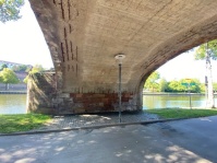 Ein Weg führt unter einer Brücke her, im Hintergrund ein Fluss