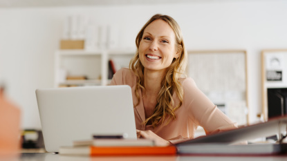Eine lachende blonde Frau sitzt an einem Schreibtisch vor einem aufgeschlagenen Laptop. Foto: contrastwerkstatt / AdobeStock