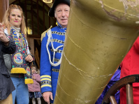 Ein Mann in blauem Kostüm steht neben einer Frau, während im Vordergrund ein großes goldenes Kanonenrohr einen Teil des Bildes verdeckt.